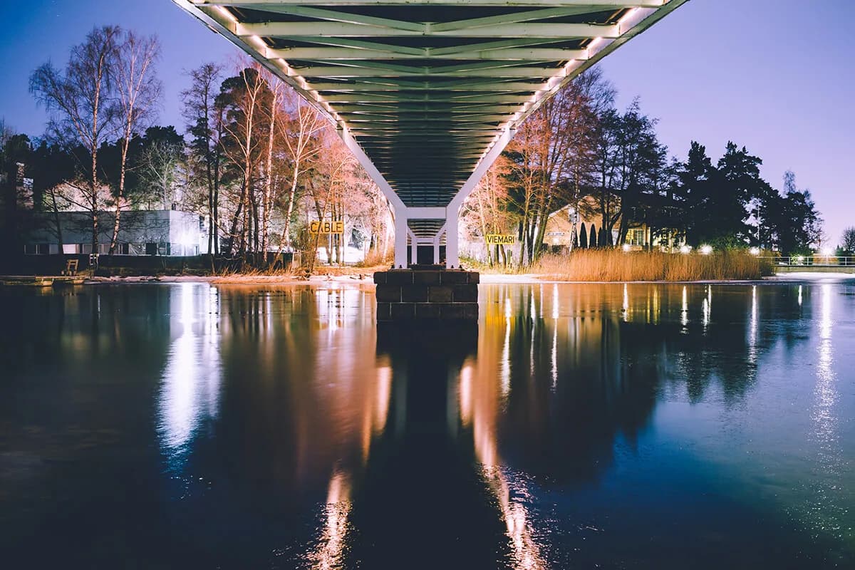 The underside of a bridge at dusk, its lights and the birch trees on both shores mirrored in still water.