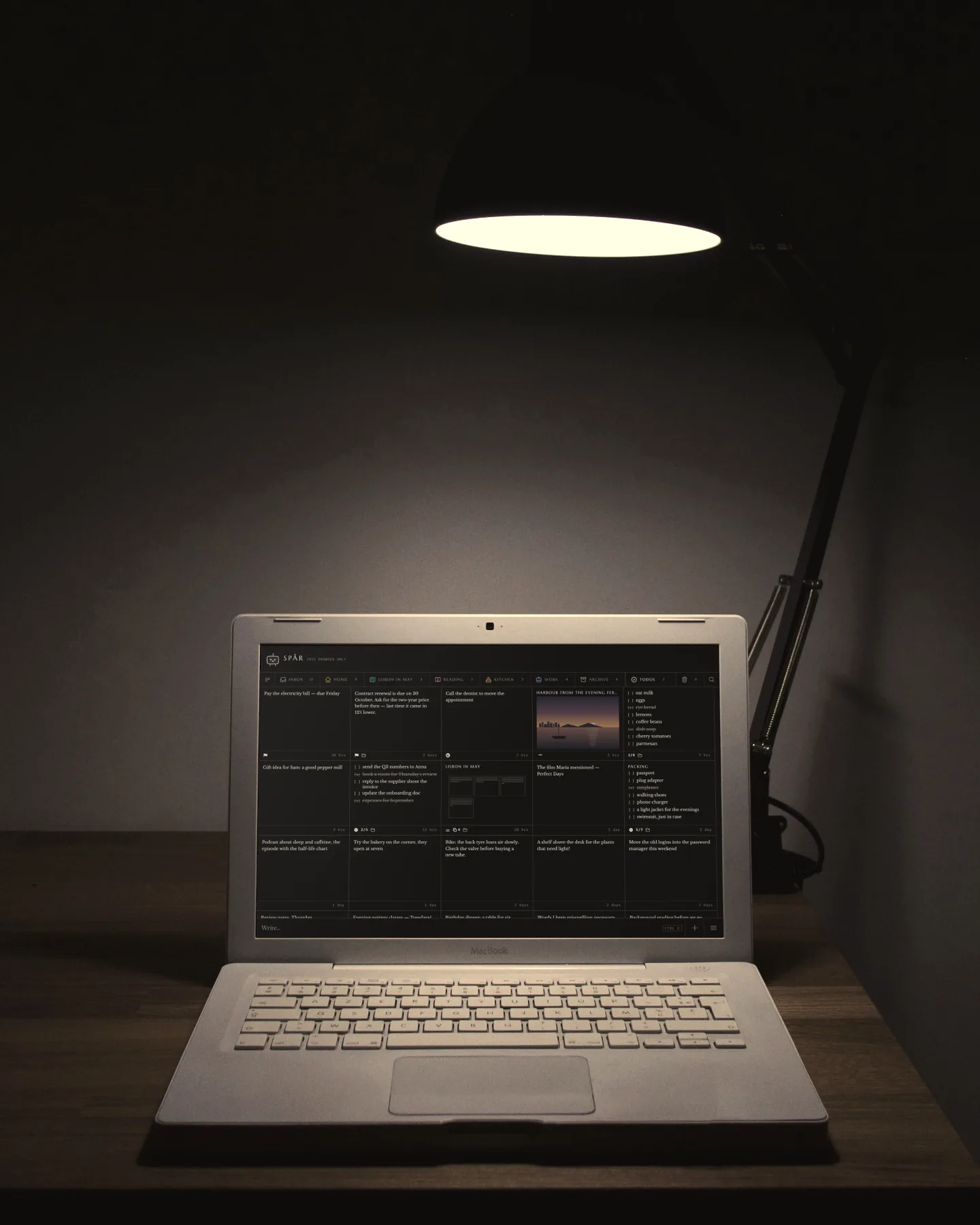 A dark room with one desk lamp: an old white laptop on a wooden desk, Spår’s notes glowing on its screen.