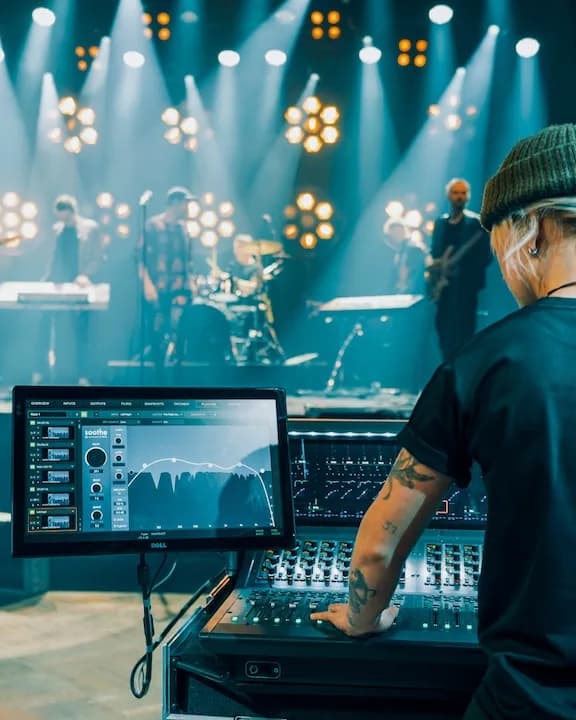 Front of house at a live show: an engineer at an Avid console, with soothe live open on the touchscreen beside her and a band on a blue-lit stage behind.