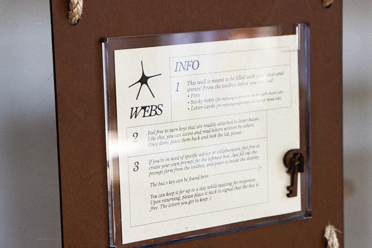 Instruction card behind clear acrylic in a brown frame, with the WEBS wordmark and spider mark on the left, three numbered steps on the right, and a laser-cut key hanging beside it.