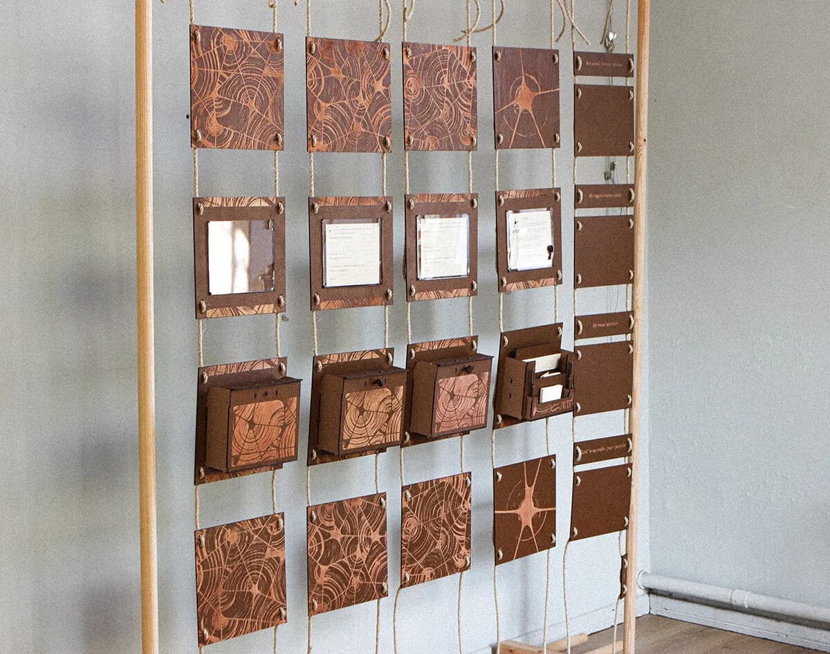 The installation in a white room: a wooden frame holding rows of brown laser-cut panels on jute rope, with engraved panels, framed prompt cards, small lockable letterboxes and a toolbox.