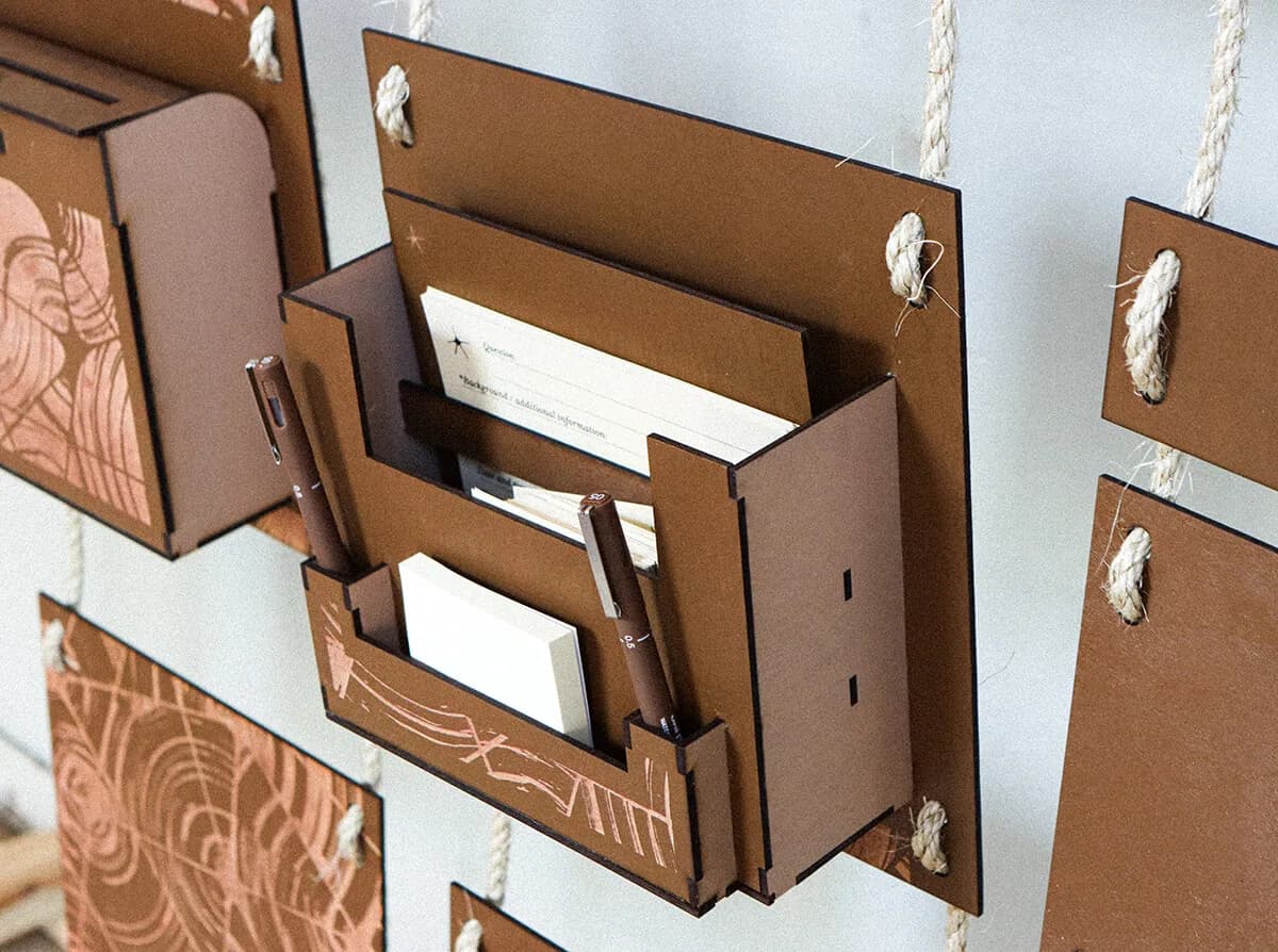 Laser-cut toolbox on the wall holding pens, sticky notes and blank prompt cards, with tab-and-slot joints visible on its sides.